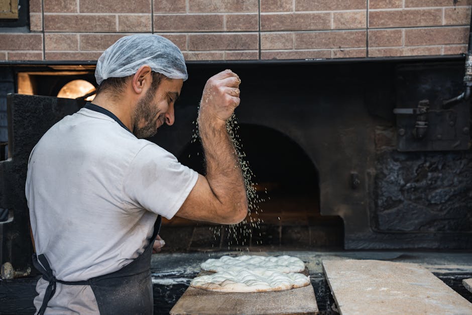 A baker skillfully sprinkles flour on flatbread dough in front of a traditional oven, showcasing baking artistry.
