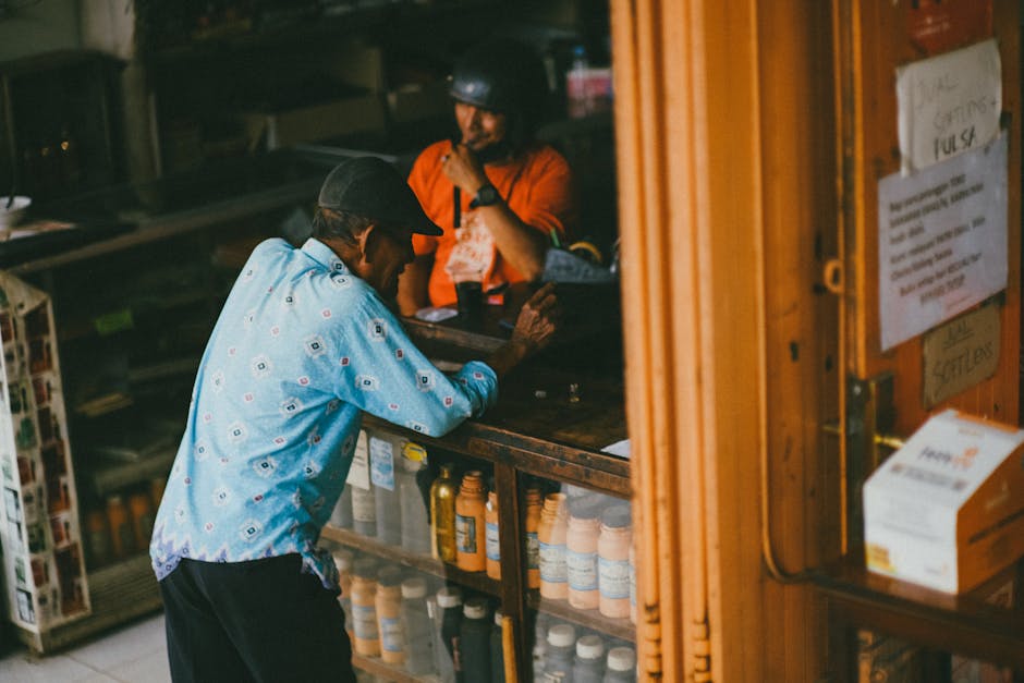 An elderly man wearing a blue shirt chats with a shopkeeper at a local store counter.